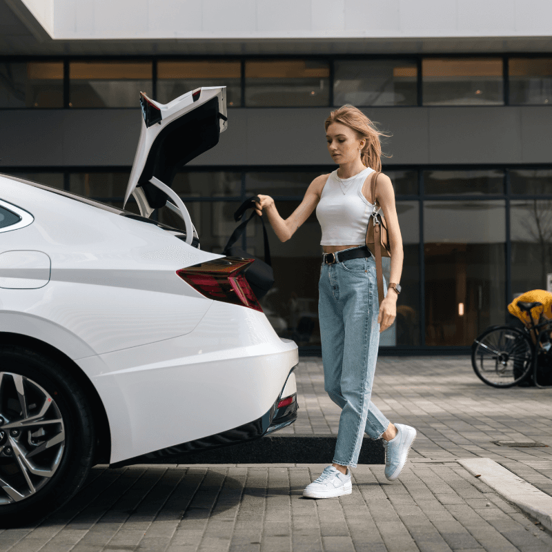 Woman loading bags into a white car trunk outside a modern building
